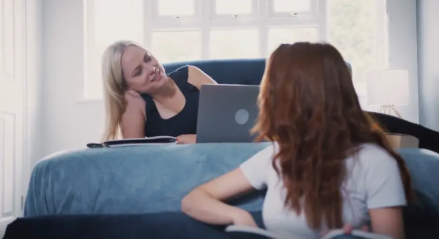 Sunlight streams into the room as two women work together on laptops, lying on a bed