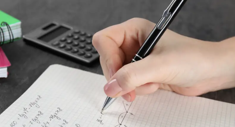 Hand writing math equations in a notebook with a calculator nearby while solving complex integrals