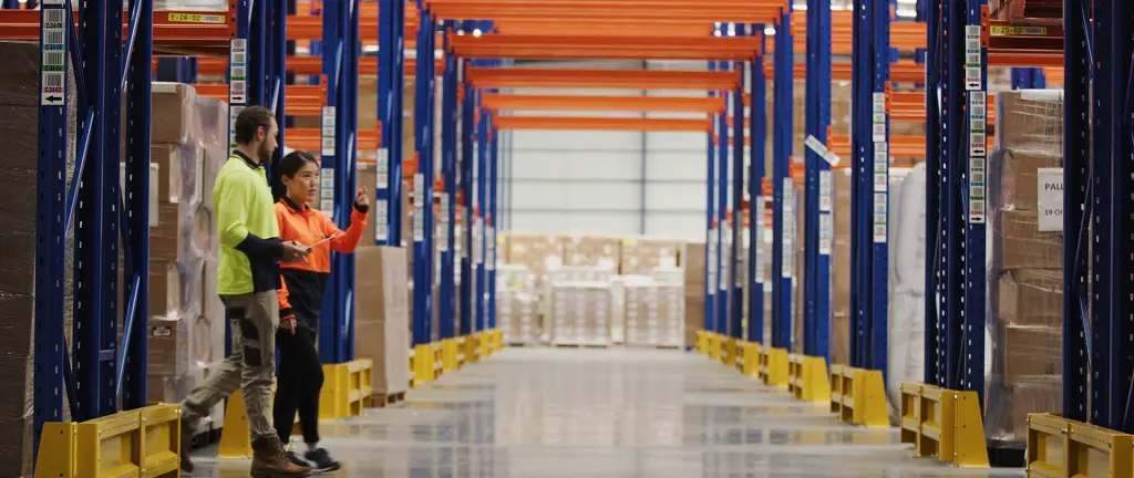 Two workers in a warehouse discussing logistics and inventory management, surrounded by tall shelving racks filled with packaged goods