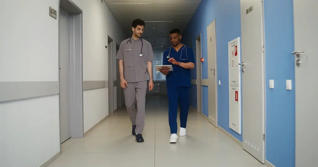 Two healthcare professionals in scrubs walking down a hospital hallway, discussing patient information on a digital tablet