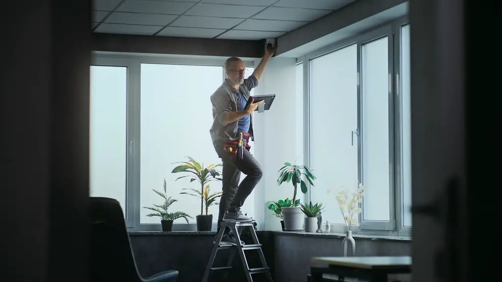 A technician on a ladder installs a security camera near a window inside an office space, holding a tablet and wearing a tool belt